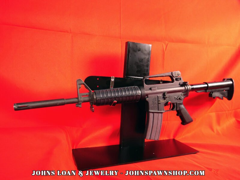 Original Bushmaster CAR-15 style rifle with a park gray finish, showcasing the firearm's profile, displayed on a black stand, available at John's Loan & Jewelry Co.