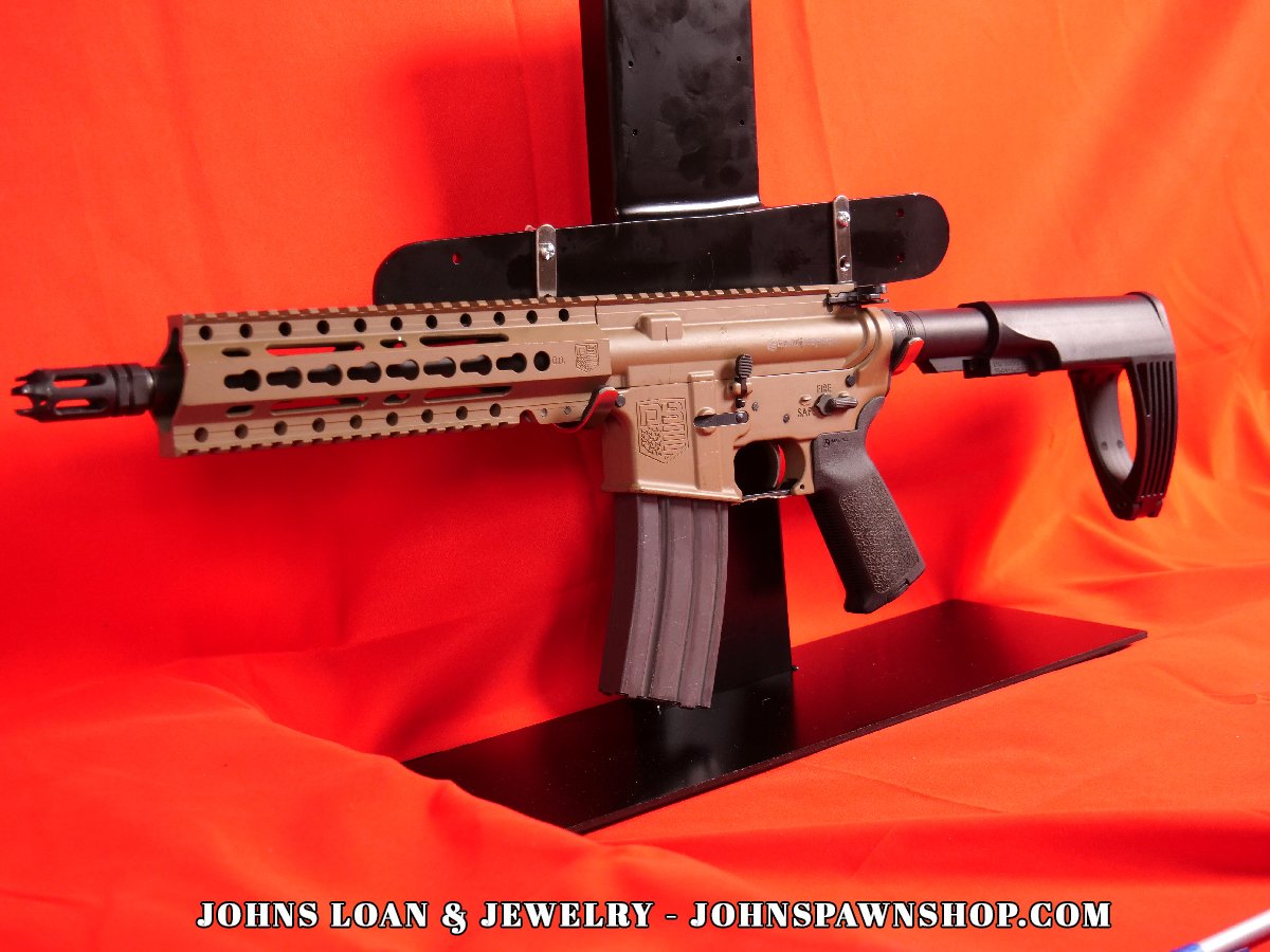 Diamondback DB15 braced AR15 pistol from John's Loan & Jewelry, displaying the firearm's FDE finish and braced design. The pistol, chambered in .223/5.56, sits on a table with a 30+1 magazine.