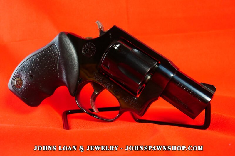 Side-profile view of a glossy black Taurus Model 85 snub nose revolver chambered in .38 Special, displayed on a red table by John's Loan & Jewelry.