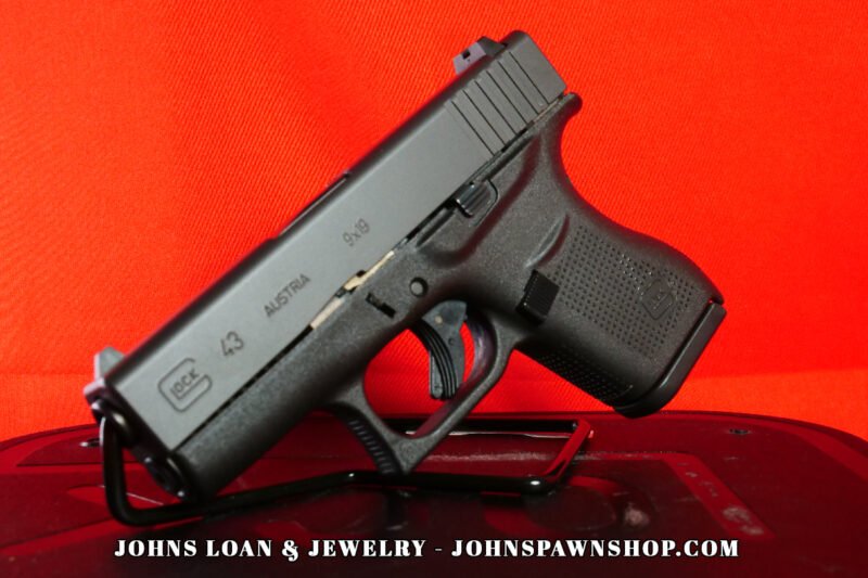 Angled view of a matte black Glock 43 semi-auto pistol in 9mm Luger, displayed on a stand; note textured grip. Available from John's Loan & Jewelry, a Las Vegas pawn shop.