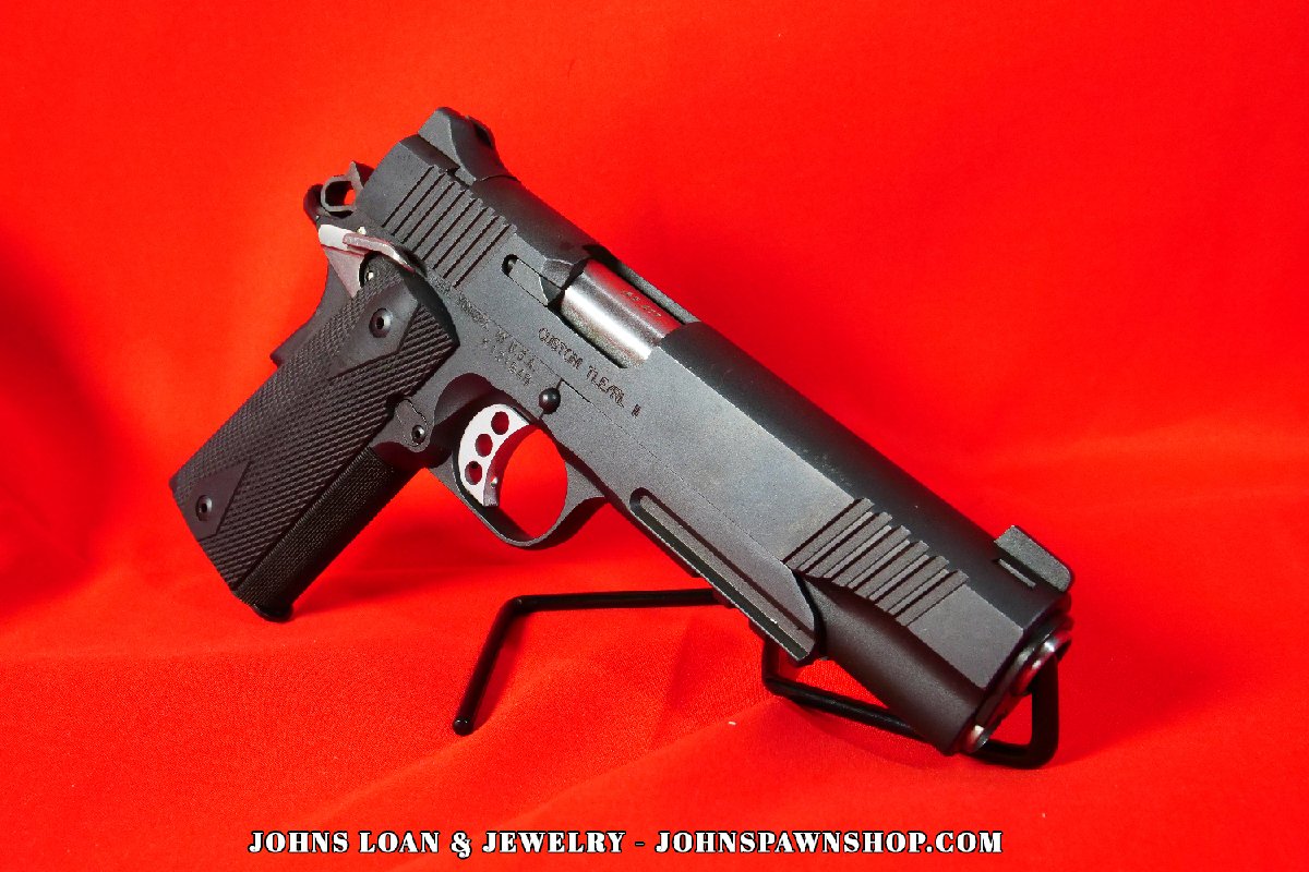 Matte black Kimber Custom TLE/RL II pistol in .45 ACP, shown on a display stand against a red backdrop, featuring diamond checkered grips, from John's Loan & Jewelry.